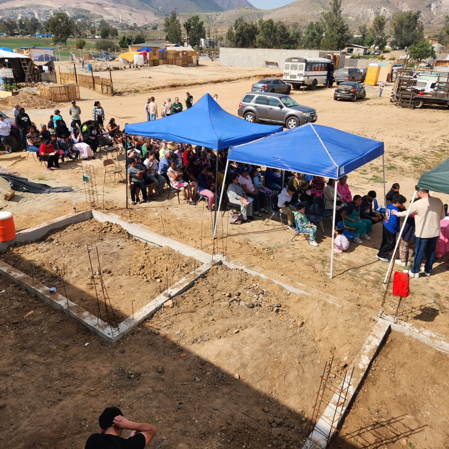 Concrete footings and rebar laid out on a hillside lot, with a crowd seated under blue canopies behind