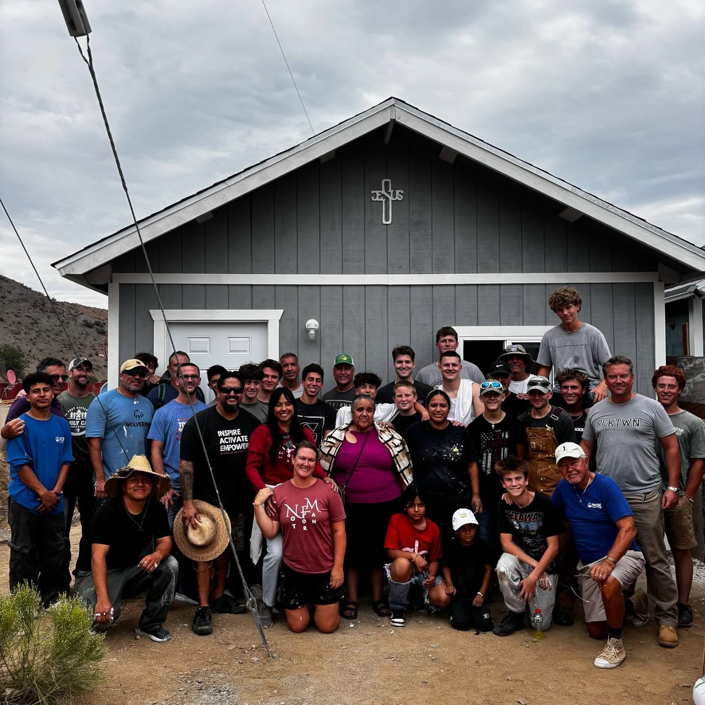 A large build team gathered in front of a finished grey house with a white cross on the gable