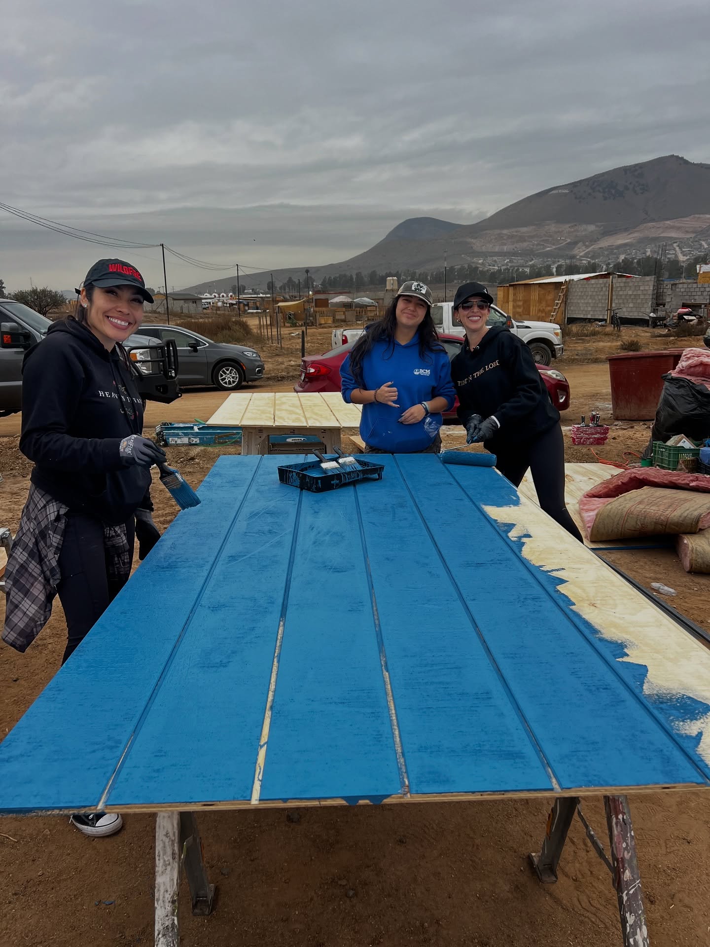 Freshly painted blue siding boards on a work table, two volunteers still rolling, with the hills behind them